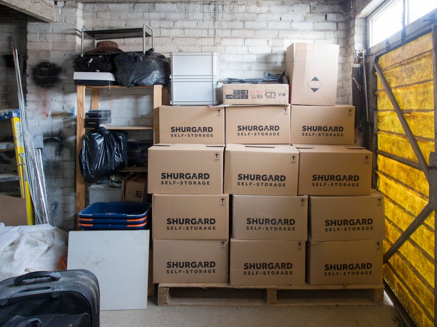 Inside a storage unit or garage with a brick wall, showing a shelving unit on the left containing black plastic bags and various smaller items. In front of the wall, there are eleven uniformly sized cardboard boxes labeled 'SHURGARD SELF-STORAGE' stacked on a wooden pallet, arranged in three rows with four boxes at the bottom, three in the middle, and four at the top. To the left of the boxes, there are stacked blue plastic trays and a white panel leaning against the boxes, with a black rolling suitcase placed nearby on the floor. On the right side, a partially visible yellow-tinted security or partition wall with metal framing is seen, suggesting an organized storage or move preparation environment. The lighting appears natural, illuminating the space and highlighting the packing and storage process relevant to home relocation or furniture transport services by Man with Van Chingford.