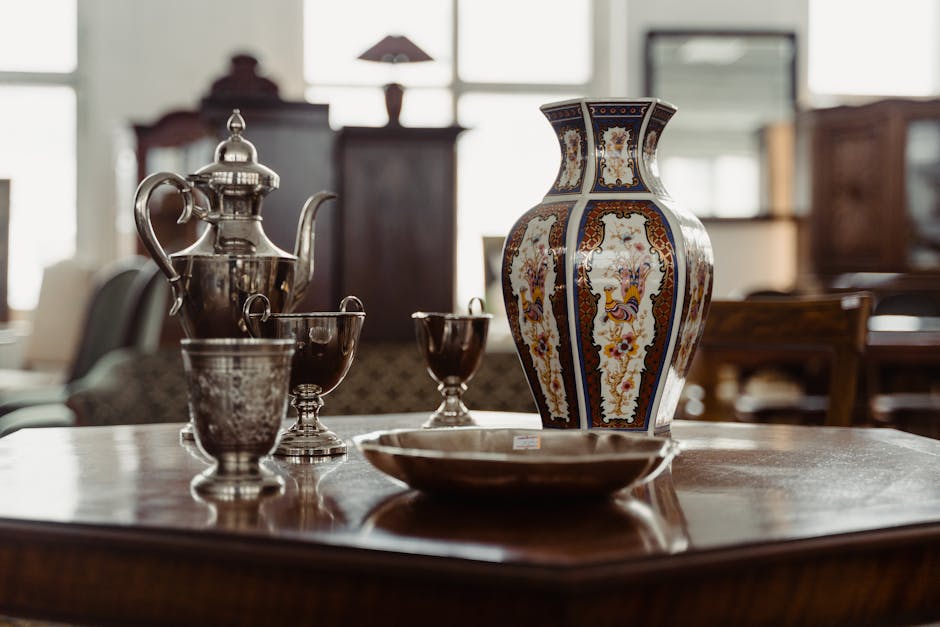 A close-up view of a wooden dining table inside a home, with a large decorative ceramic vase featuring intricate floral and geometric patterns centrally positioned on the table. Surrounding the vase are various silverware pieces, including a teapot with a curved spout and handle, a matching sugar bowl, and several goblets or cups, all arranged neatly. A large shallow bowl is placed in front of the vase, possibly for serving or packing purposes. The background shows a blurred interior with furniture, including a sideboard or cabinet, soft natural light coming through windows, and a cozy, well-lit environment. This image captures a moment during packing or moving preparations, where items might be being organized for transport, linked to home relocation services, as provided by Man with Van Chingford.