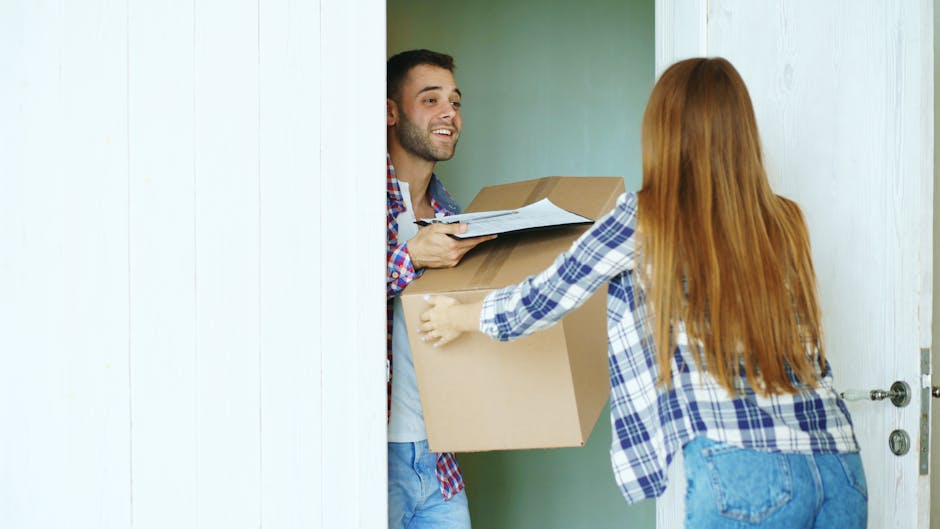 A man with short dark hair and a beard, wearing a blue and red checked shirt over a white T-shirt, stands inside a doorway holding a cardboard box packed with household items, while a woman with long red hair, dressed in a blue and white checked shirt and jeans, reaches out to hand the man a package or document. The scene takes place at the entrance of a property, with the door open, revealing a light-colored interior wall. There is a focus on furniture and packing materials, including the cardboard box, which appears to contain books or packages, and possibly additional boxes stacked behind or around them. The setting indicates a home relocation or moving process, with the manwithvanchingford.co.uk's removals services likely involved in transporting household belongings. The lighting is natural, emphasizing clarity and professionalism during the packing and loading stages of house removals.
