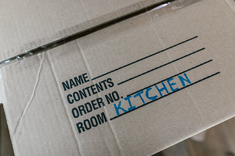Close-up of a cardboard moving box within a home, showing a label with the words 'NAME', 'CONTENTS', 'ORDER NO.', and 'ROOM', handwritten in black and blue ink, with 'KITCHEN' written in blue. The box is made of brown corrugated cardboard, partially wrapped in clear plastic, and positioned on a wooden floor near a doorway. In the background, the interior of a property is visible, with natural light illuminating the area, indicating an active home relocation or packing process carried out by Man with Van Chingford, a professional removals service specializing in house moves and furniture transport.