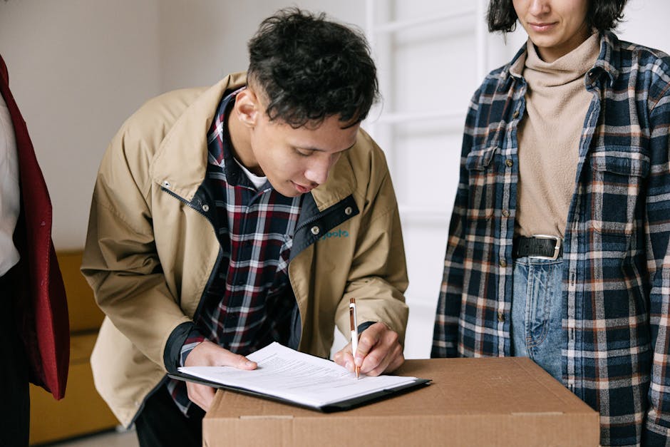 A man with short dark hair and a beard, wearing a blue and red checked shirt over a white T-shirt, stands inside a doorway holding a cardboard box packed with household items, while a woman with long red hair, dressed in a blue and white checked shirt and jeans, reaches out to hand the man a package or document. The scene takes place at the entrance of a property, with the door open, revealing a light-colored interior wall. There is a focus on furniture and packing materials, including the cardboard box, which appears to contain books or packages, and possibly additional boxes stacked behind or around them. The setting indicates a home relocation or moving process, with the manwithvanchingford.co.uk's removals services likely involved in transporting household belongings. The lighting is natural, emphasizing clarity and professionalism during the packing and loading stages of house removals.