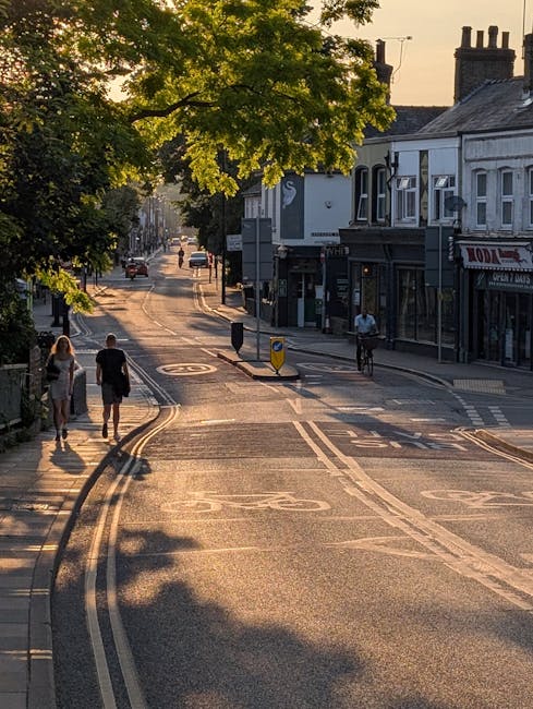 A quiet residential street scene during late afternoon or early evening, with a large leafy tree casting shadows over the pavement and road. On the left side, three pedestrians—two women and one man—are walking along the sidewalk, while on the right, a cyclist rides on the roadway. The street is lined with traditional brick and painted shopfront buildings, some with visible signage, and features a set of double yellow lines along the curb. In the distance, a few cars are visible traveling down the road, which curves gently ahead, illuminated by warm sunlight. The overall scene depicts a peaceful urban environment suitable for house removals or home relocation services, with the street’s layout and nearby shops providing context for logistics and transportation activities by companies like Man with Van Chingford.