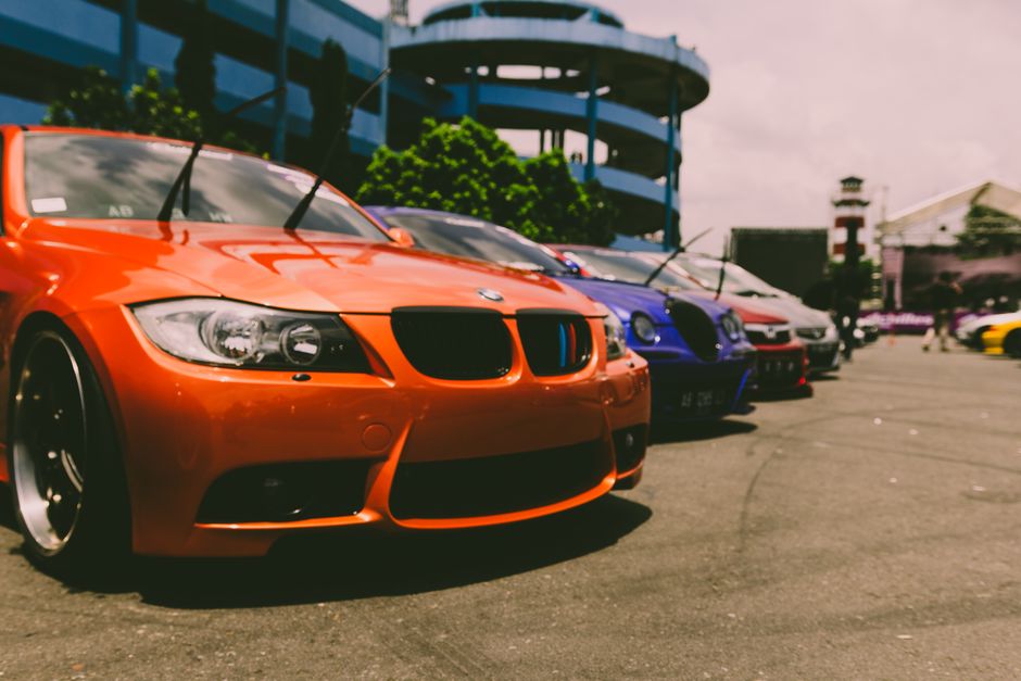 A row of parked cars on the street includes a bright orange sports car with a sleek design and black accents, positioned closest to the camera. Behind it, there is a blue car and several other vehicles, all lined up parallel to the pavement. The cars are parked near a building with a modern, curved blue balcony structure, and some greenery is visible in the background. The scene appears to be outdoors during daytime, with natural light illuminating the vehicles and surroundings. Occasionally, a person can be seen walking in the distance, and the environment suggests an urban area potentially associated with house removals or furniture transport services. Man with Van Chingford occasionally operates in such areas involving logistics, packing, and vehicle loading, aligning with the theme of house relocations and transportation logistics.