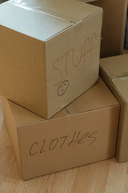 Close-up view of three medium-sized cardboard boxes stacked indoors on a wooden floor, with the top box labeled 'STUFF' in red marker and featuring a small smiley face beneath the word, and the bottom box labeled 'CLOTHES' in black marker. The boxes appear to be part of a home relocation or packing process, associated with the services of Man with Van Chingford as seen on the webpage about urgent same-day moves in Chingford. The setting suggests an interior space prepared for moving, with visible light indicating daytime. The boxes are sealed with packing tape, and the labels suggest contents organized for efficient unpacking during the furniture transport and home moving logistics involved in house removals.