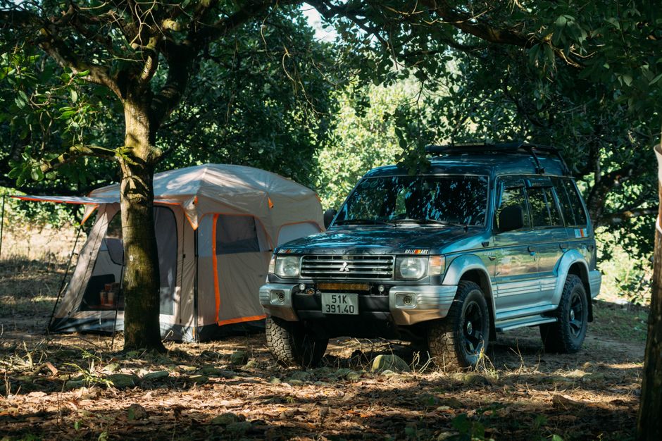A blue Mitsubishi four-wheel-drive vehicle with black wheels and a roof rack is parked on a patch of dirt and grass amidst trees with dense green foliage. Nearby, there is a beige and orange camping tent made of fabric with mesh windows and a zippered door, partially shaded by the trees. The tent appears to be set up in a natural outdoor area, possibly for a home relocation or temporary accommodation during a move. The scene is lit by natural daylight, with sunlight filtering through the leaves, creating dappled shadows on the ground and on the vehicle and tent. The setting suggests a campsite or outdoor staging area, with the vehicle positioned close to the tent, indicating a practical moving or packing process in a woodland environment, consistent with relocation services like those offered by Man with Van Chingford.