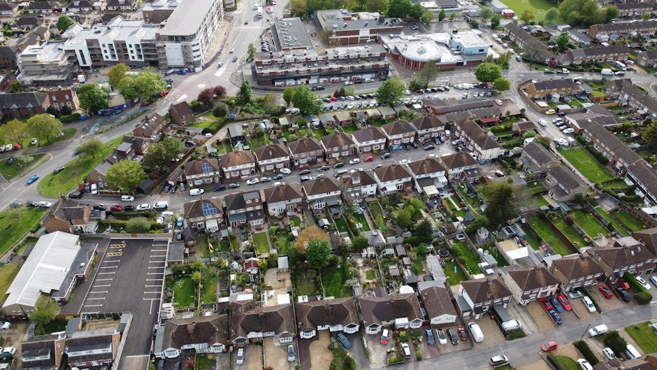 Aerial view of a residential neighbourhood during daytime showing closely spaced rows of terraced houses with small front gardens and driveways, some with parked cars. The houses are predominantly brick-built with tiled roofs, and many have solar panels installed on their roofs. Behind the row of terraced houses, there is a parking lot filled with vehicles, and beyond that, larger commercial or apartment buildings with modern white facades. The area is lush with green trees and landscaped gardens, indicating a well-maintained suburban environment. This setting provides context for home relocation or furniture transport activities, with visible signs of moving logistics such as packed boxes or furniture possibly being carried in or out of properties, although specific items are not clearly shown. Man with Van Chingford likely uses such locations for moving services, including packing, loading, and transport within the neighbourhood as part of household removals or relocation services.